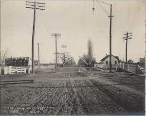 City of Toronto Archives, c. 1900, Bloor St looking east across rail tracks towards Lansdowne Ave. The house in the photo is in the area of the Lochrie residence and adjacent rope and bicycle-making factory. A path is visible on the north side of the road beside the sidewalk.